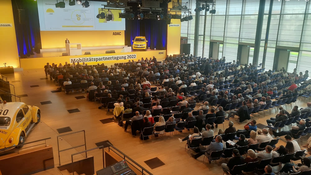 Vollbesetzter Saal einer Mobilitätspartnertagung mit Bühne, Leinwand und Fahrzeug